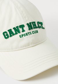 Khaki baseball cap with green embroidered text "GANT NHCT. SPORTS CLUB." Features curved visor and stitched detailing on the fabric.