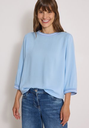 Femme souriante aux cheveux bruns portant un haut ample bleu clair à manches longues et un jean bleu, debout devant un fond uni.