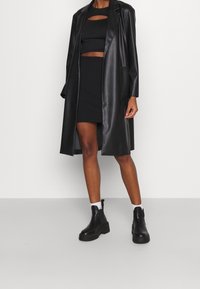Cappotto lungo in ecopelle nera, sovrapposto a un top corto nero con aperture e una gonna nera aderente. Un paio di stivali alla caviglia neri chunky.