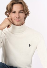 Maglione bianco a collo alto in tessuto morbido, con colletto a coste e un piccolo logo scuro sul lato sinistro del petto, indossato con jeans in denim.
