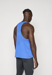 Niebieski sportowy top na ramiączkach z siateczkowym wzorem, otwartym plecami i delikatną teksturą. Noszony z dopasowanymi czarnymi legginsami z białym logo.