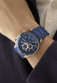 Mėlynas chronografas laikrodis su rožių aukso rėmeliu, guminiu diržu ir detaliu ciferblatu, kuris turi subciferblatus ir švytinčius žymeklius, uždėtas ant riešo.