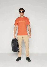 Oransje kortermet T-skjorte med Columbia-logo, beige bukser, svart ryggsekk og solbriller. Person står foran en nøytral bakgrunn.