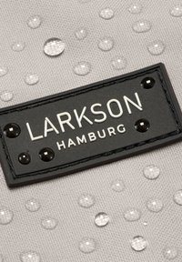 Közelkép egy fekete, téglalap alakú címkéről, rajta a "LARKSON HAMBURG" felirattal, amely világos anyagból készült és vízcseppekkel van borítva.