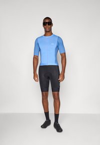 Modrá cyklistická dres s krátkým rukávem a předním zipem, doplněný černými cyklistickými kraťasy. Model má na sobě černé cyklistické boty a sluneční brýle.