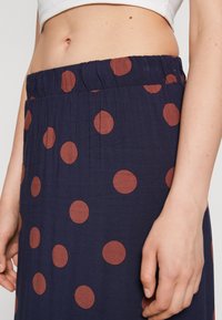 Falda de color azul marino con cintura fruncida, que presenta grandes lunares marrones. Tejido ligero con una textura suave. Mano visible descansando sobre la tela.
