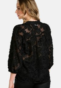 Black sheer blouse with textured floral embroidery, featuring three-quarter sleeves and a rounded neckline.  Soft fabric with visible stitching.