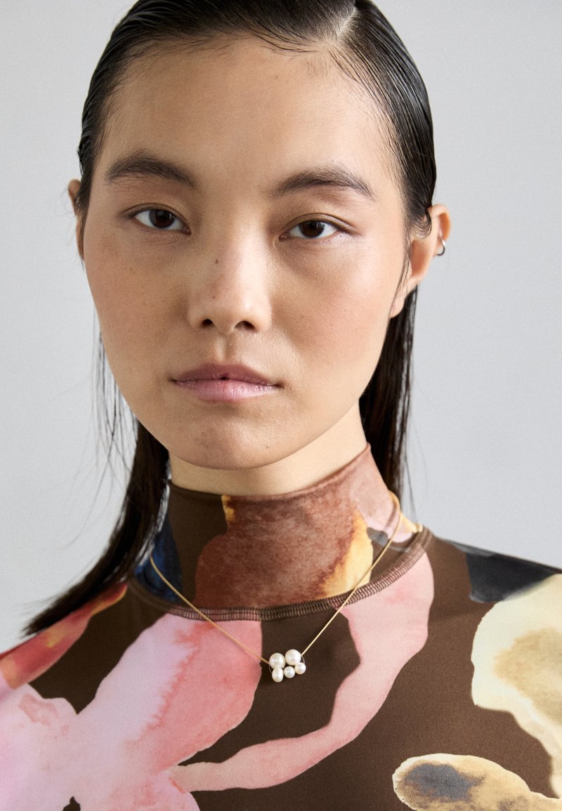 Τοπ με ψηλό λαιμό και ανθισμένο σχέδιο σε καφέ, ροζ και μπλε αποχρώσεις. Χρυσό κολιέ με τρεις ομάδες λευκών μαργαριταριών γύρω από το λαιμό.