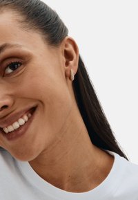 Kolczyki na sztyft w kolorze złotym, o kształcie kropli i fakturowanej powierzchni, noszone przez kobietę z długimi włosami, delikatnie się uśmiechającą.