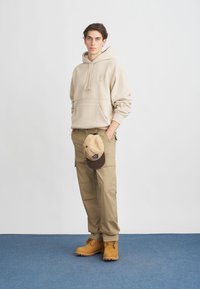 Hanorac bej din fleece cu buzunar frontal, asortat cu pantaloni cargo khaki. Accesoriile includ o șapcă bej și cizme bej rezistente. Design simplu și funcțional.