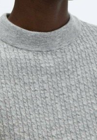 Persona di carnagione scura che indossa un maglione grigio chiaro a maglia strutturata con un colletto a coste.
