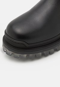 Botte en cuir noir avec un bout arrondi et une semelle épaisse et translucide à crampons profonds visible sur fond blanc.