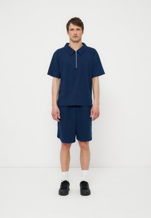 Man standing against white wall wearing navy short-sleeve zip-neck shirt, matching shorts, white socks, and black sneakers.