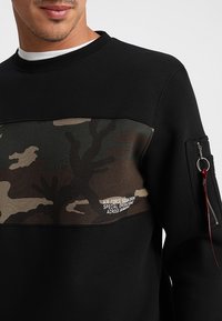 Felpa nera con un pannello camouflage e tasca con zip sulla manica. Dettagli testuali sulla sezione camouflage. Collo a costine, tessuto testurizzato.