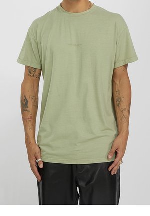 Uomo che indossa una maglietta verde chiaro a maniche corte e pantaloni neri, mostrando tatuaggi su entrambi gli avambracci e anelli alle dita.