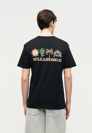 Moški nosi črno majico Billabong z risbami pastelnega sonca, rastline in palme na hrbtni strani, združeno s svetlo sivimi kratkimi hlačami.