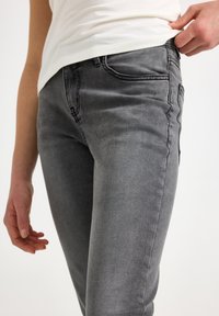 Άτομο που φοράει εφαρμοστά γκρι τζιν και λευκό πουκάμισο, με το ένα χέρι να ρυθμίζει τη μέση.