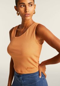 Top corto sin mangas de color beige con canalé que cuenta con un amplio escote redondeado y un logo bordado en el dobladillo. Combinado con una tela azul de textura suave.