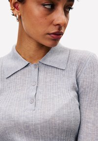 Vrouw met kort haar, die zilveren en gouden ringoorbellen draagt, en een lichtgrijze geribde trui met kraag en knopen.