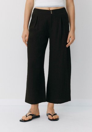 Femme portant un pantalon noir taille haute à jambes larges avec des plis à l'avant et deux boutons, assorti de sandales noires et de tatouages visibles sur les pieds.