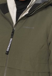 Olijfgroen pufferjack met een ritssluiting aan de voorkant, voorzien van een zachte binnenvoering, afneembare capuchon en een merkrits.