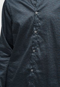 Σκούρο μπλε πουκάμισο με ανοιχτό μπλε γεωμετρικό μοτίβο "FX" και μαύρα κουμπιά μπροστά, φορεμένο με τα μανίκια κατεβασμένα.