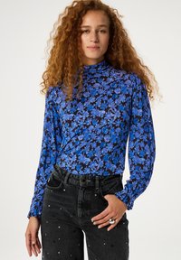 Langärmliges Oberteil mit hohem Kragen, versehen mit einem blauen Blumenmuster auf schwarzem Hintergrund. Kombiniert mit hoch taillierten, dunklen Jeans, die mit Perlen verziert sind.