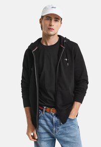 Schwarze Zip-Hoodie mit strukturierter Oberfläche, darunter ein weißes T-Shirt, dazu Jeans und eine weiße Cap. Schlichtes Design mit dezenten Details.