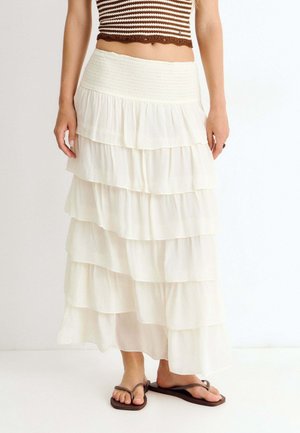 Femme portant une jupe maxi blanche à plusieurs étages et un top court rayé marron, debout pieds nus sur un sol blanc contre un mur uni.