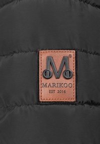Svart puffjakke med et fremtredende brunt skinnmerke med preget logo "M" og teksten "MARIKOO EST 2016," sydd til stoffet.