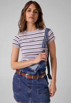 Jeune femme portant un t-shirt rayé Guess Jeans et une jupe salopette en denim avec une bretelle détachée, sur un fond uni.