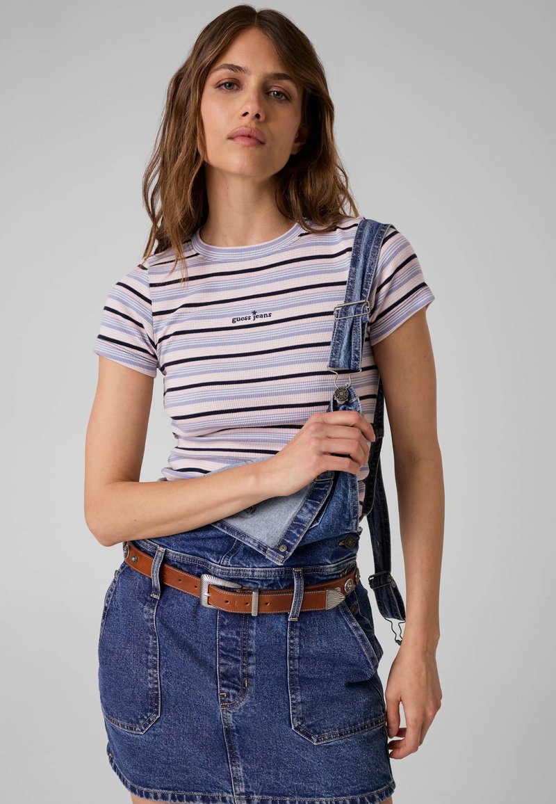 Νεαρή γυναίκα που φοράει ριγέ μπλουζάκι Guess Jeans και τζιν σαλοπέτα φούστα με ένα λουρί χαλαρό, μπροστά σε απλό φόντο.