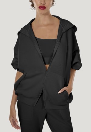 Vrouw die een oversized zwarte capuchontrui met rits draagt, een zwart cropped topje en bijpassende zwarte broek, met één hand in de zak, tegen een effen achtergrond.