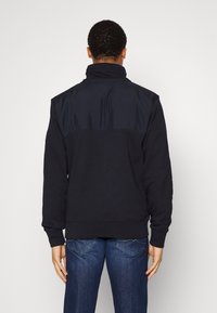 Sudadera negra con cuello alto, que presenta un panel de tela brillante en los hombros. Textura suave con dobladillo y puños acanalados.