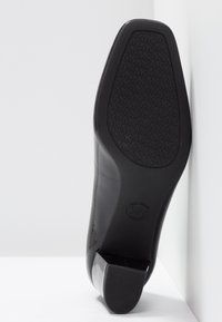 Semelle de chaussure de ville noire avec un grip à motif comprenant les initiales répétées « MK » et un talon carré, inclinée contre un fond blanc en coin.