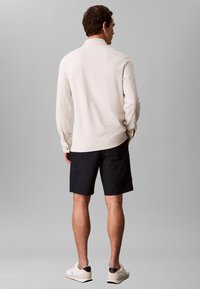 Cămașă lungă cu mânecă lungă, bej deschis, cu guler, asortată cu pantaloni scurți negri și adidași albi cu detalii închise la călcâi.