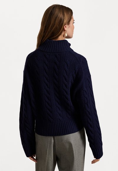 Navy blue cable-knit sweater with a high collar, loose fit, and ribbed hem. The fabric appears thick and textured, showcasing detailed patterns.