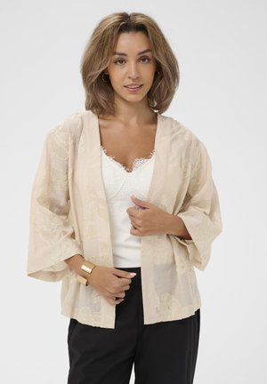 Kobieta o włosach długości do ramion w jasnobrązowym kolorze, ubrana w beżowe kwiatowe kimono na białej koronkowej bluzce i czarne spodnie, z złotą bransoletą na nadgarstku.