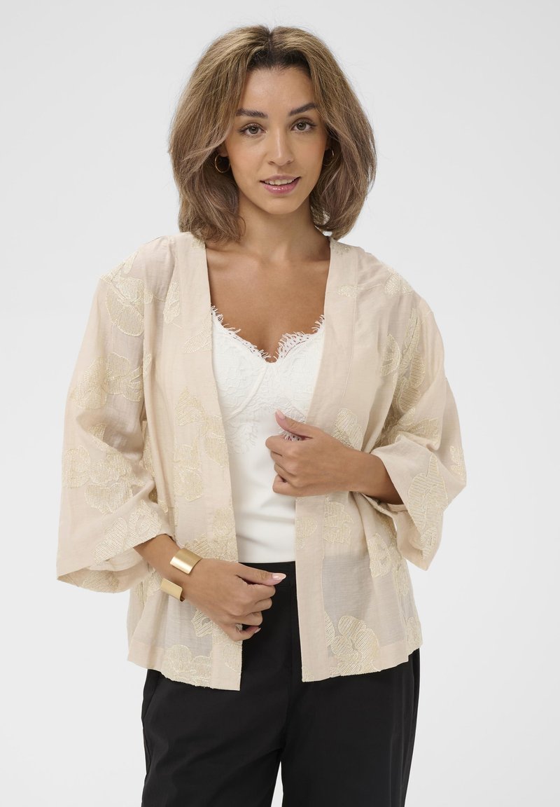 Vrouw met halflang lichtbruin haar, gekleed in een beige bloemenkimono over een witte kanten top en zwarte broek, met een gouden manchetarmband.