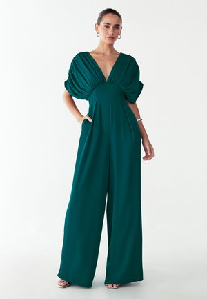 Turkis buksedragt med vide ben, dyb V-udskæring og pufærmer. Fremstillet af glat stof med en tætsiddende talje og sidelommer.