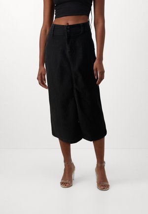 Kvinde iført sort ærmeløs crop top, sort højtaljet midinederdel og klare stropper med højhælede sandaler står på en hvid gulv.
