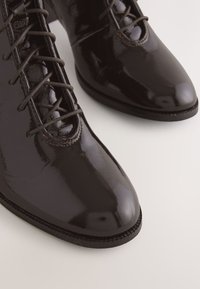 Sorte ankelstøvler i laklæder med fint tekstureret overflade, snørestil, spids tå og kontrastsyninger. Glat, blank finish.