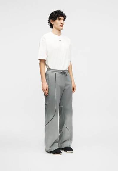 Άνδρας με σγουρά μαλλιά, που φοράει λευκό μπλουζάκι με κοντά μανίκια και άνετο ανοιχτό γκρι παντελόνι με μαύρα σνίκερς, στέκεται μπροστά σε ένα απλό φόντο.