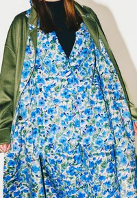 Robe à motif floral aux tons bleus et verts avec un profond décolleté en V, portée sous un manteau vert olive doté de gros boutons et de poches.