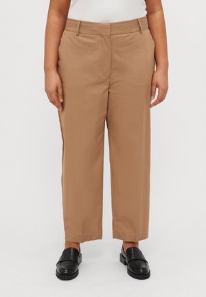 Person iført beige bukser med lige ben og sorte slip-on sko står foran en ensfarvet hvid baggrund.