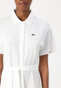 Balta polo suknelė su trumpomis rankovėmis, knopkių uždarymu ir rišamu liemeniu. Ant kairiojo krūtinės puikuojasi mažas žalias Lacoste logotipas. Minkšta tekstūra.