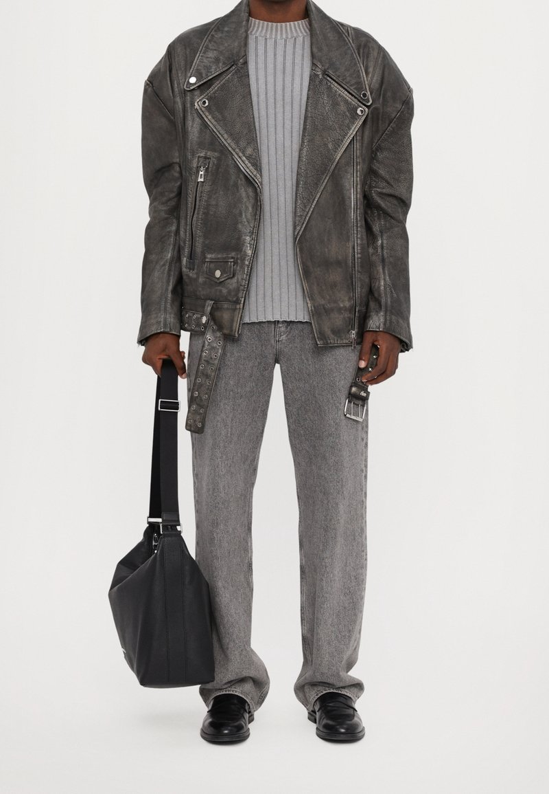Homme portant une veste en cuir usée surdimensionnée, un pull gris côtelé, un jean gris ample, des chaussures noires, tenant un sac à bandoulière noir.