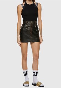 Top sans manches noir avec des bretelles logo, associé à une mini-jupe en cuir noir taille haute avec des fermetures éclair et des poches, stylisé avec des chaussettes blanches et des claquettes.