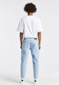 Persona con cabello rizado vestida con una camiseta blanca, jeans de mezclilla azul claro y zapatillas blancas, de pie frente a un fondo blanco liso.