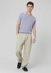Polo en maille lavande avec col, associé à un pantalon large beige clair et des mocassins noirs, se tenant devant un arrière-plan neutre.